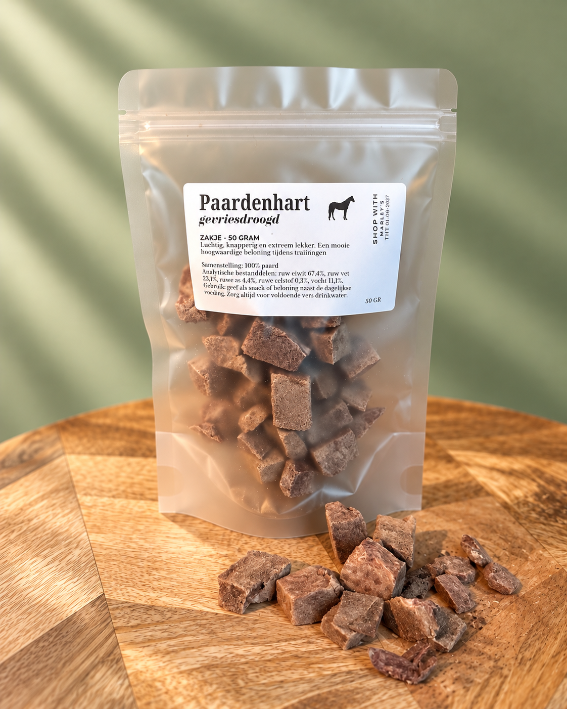 Marley’s gevriesdroogd paardenhart 50 gram in hersluitbaar zakje, met losse stukjes paardenhart op houten plateau en zachtgroene achtergrond