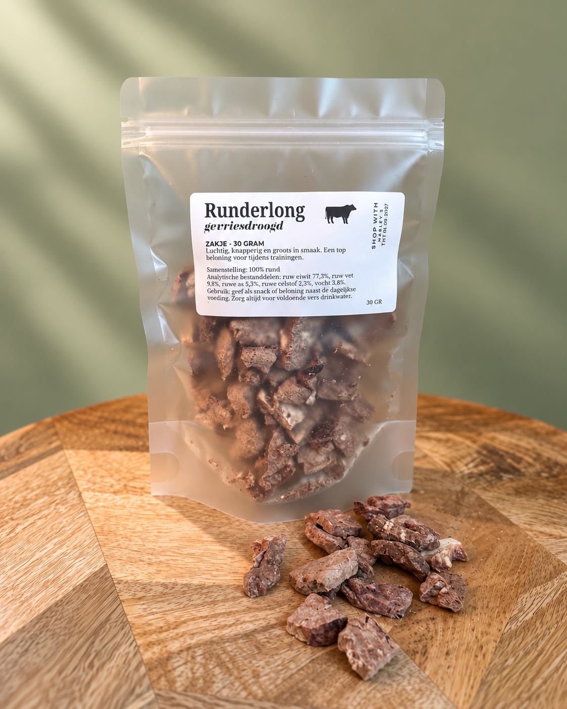 Marley’s gevriesdroogde runderlong 30 gram in hersluitbaar zakje op houten plateau met losse runderlongsnacks voorop tegen een zachtgroene achtergrond